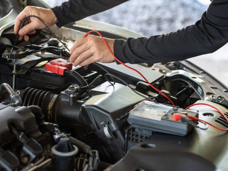 A technician provides Battery Services by using a multimeter to check voltage on a car battery under the hood of a vehicle.
