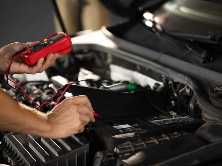A person uses a digital multimeter to test a car battery under the open hood of a vehicle, checking its health as part of routine battery services.
