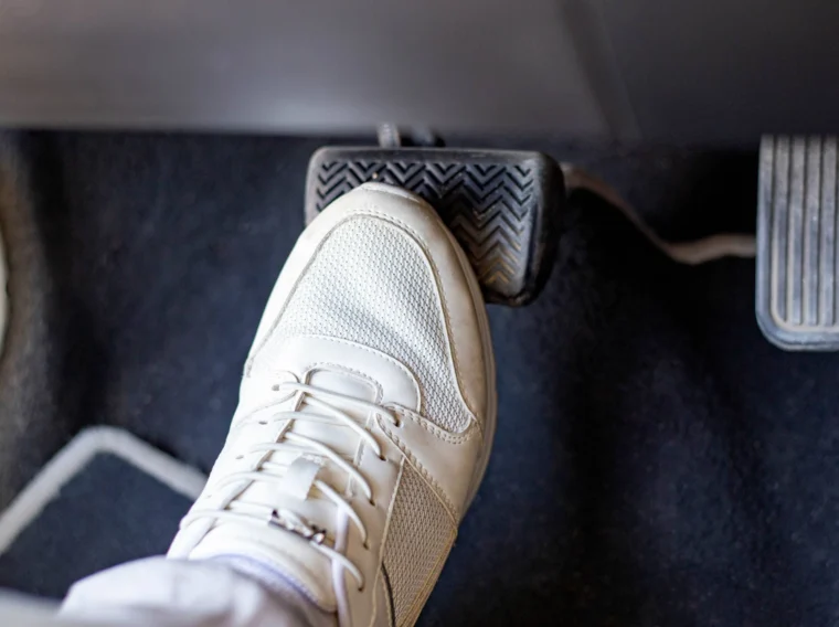 A close-up of a foot in a white sneaker pressing down on a car's brake pedal, next to the accelerator, highlights the importance of proper clutch replacement for smooth driving on a dark car floor.