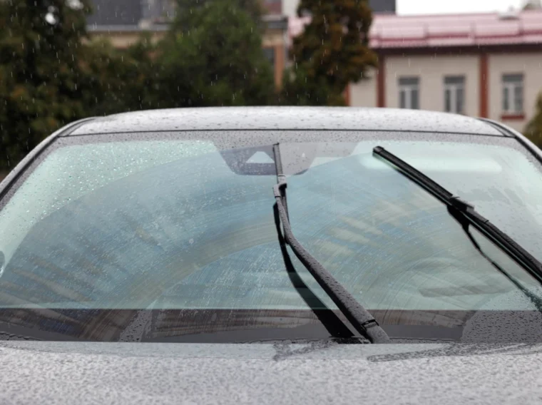 A close-up of a car windshield with two windshield wipers in motion, removing rainwater as it rains, with buildings and blurred lights visible in the background.
