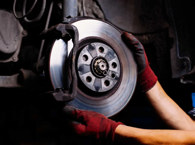 Mechanic wearing red gloves working on a car’s exposed brake disc and caliper, performing maintenance or repair as part of professional brake services.