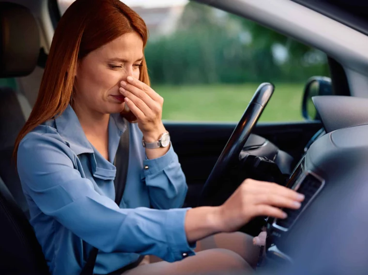 A woman sitting in the driver's seat of a car holds her nose while adjusting controls on the dashboard, possibly indicating a need for cylinder replacement due to an unpleasant odor inside the vehicle.