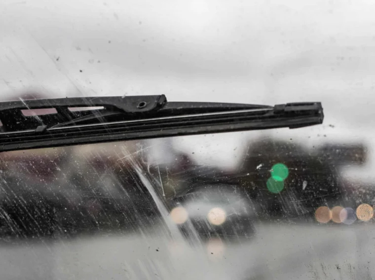A windshield wiper sweeps across a dirty, streaked windshield on a rainy day, with blurred lights and traffic signals glowing in the background.