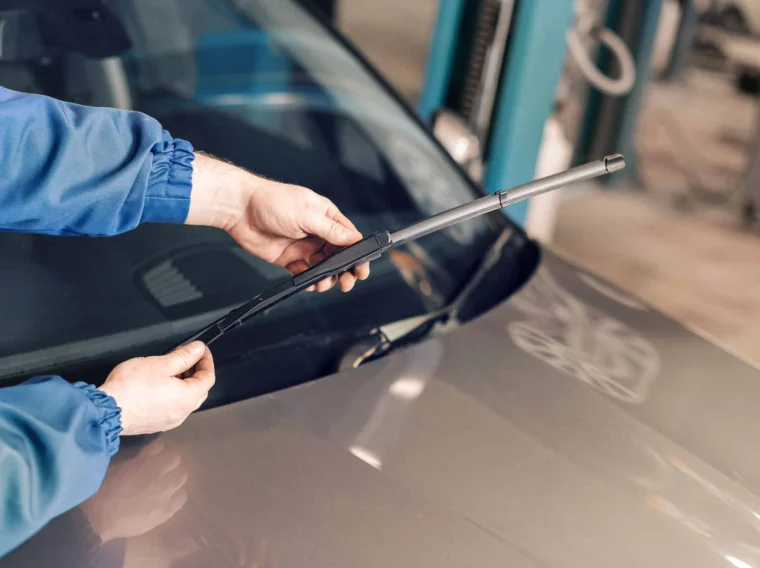 Person in blue sleeves installing a windshield wiper blade on a car in a garage, ensuring the windshield wipers and lights are well-maintained for safe driving.
