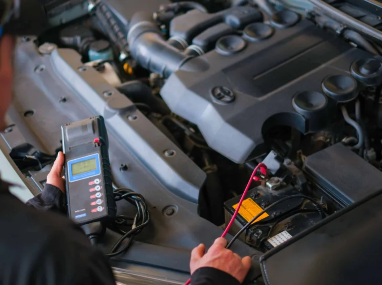 A person uses a diagnostic tool to check the battery of a car with its hood open in a workshop, ensuring reliable Battery Services.