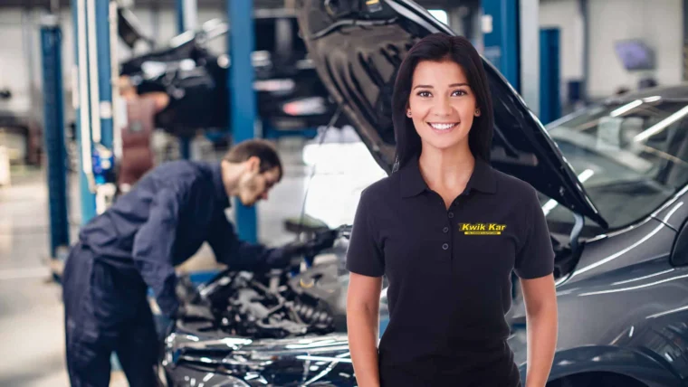 A woman in a Kwik Kar uniform stands smiling in an auto repair shop while a mechanic works on a car in the background.