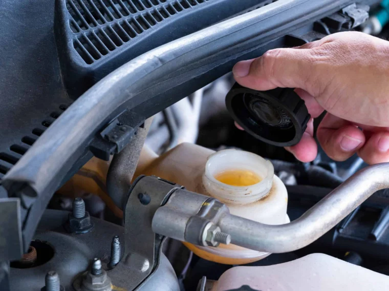 A hand opening the brake fluid reservoir cap under a car hood, with brake fluid visible inside the container.