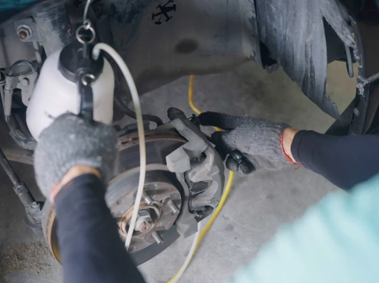 Person wearing gloves uses a brake bleeder tool to service the car’s brake system with the wheel removed, exposing the brake rotor and caliper.