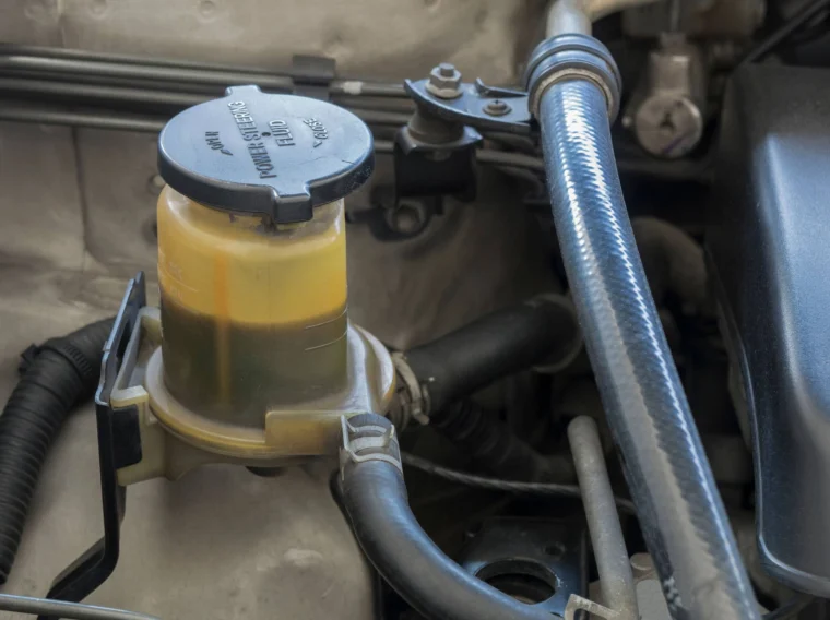 Close-up view of a car engine bay showing a brake fluid reservoir with dirty, discolored fluid and visible contamination near the power steering components.
