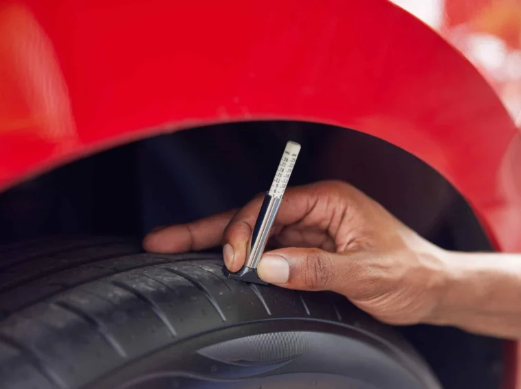 A person measures the tread depth of a car tire using a tread depth gauge under the wheel arch of a red vehicle as part of routine Tire Services.