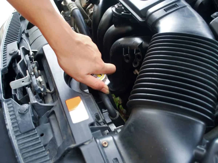 A hand is opening a car radiator cap under the hood, with various engine components visible, demonstrating the importance of Fluid Exchange Services.
