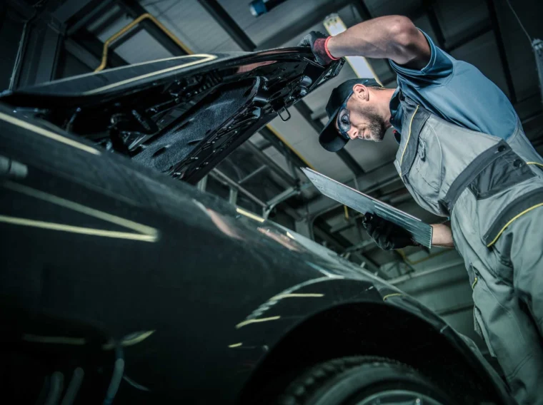A mechanic in a workshop uniform inspects a car engine under an open hood while holding a laptop computer, showcasing professional Vehicle Inspection services.