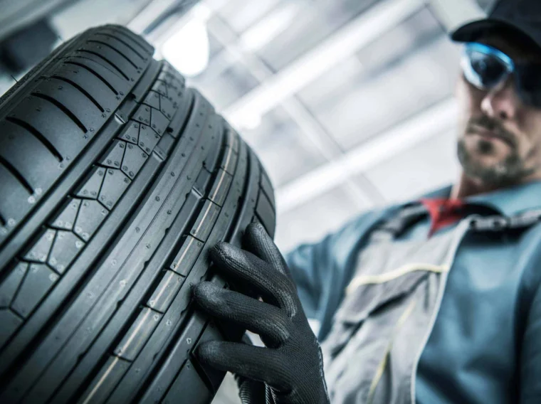 A mechanic wearing gloves and safety glasses inspects the tread of a car tire in a well-lit workshop, ensuring top-quality Tire Services for safe driving.