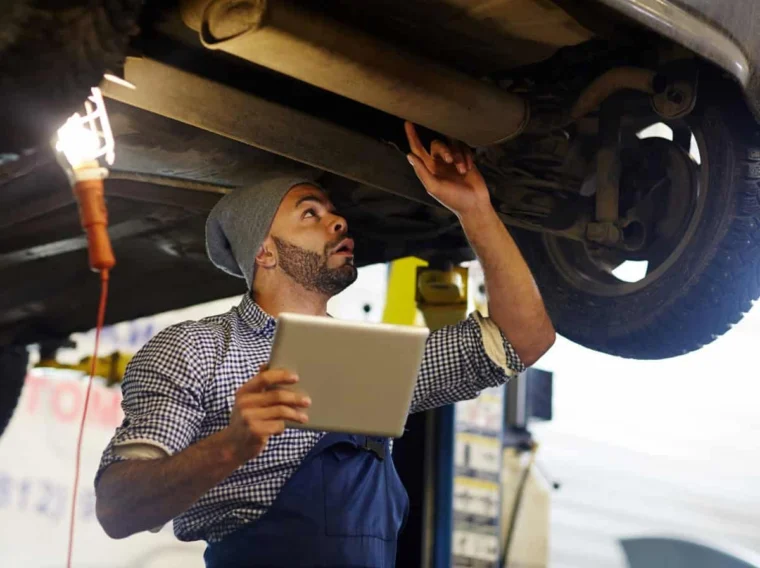 A mechanic wearing a beanie inspects the underside of a car while holding a tablet, showcasing thorough Vehicle Inspection services, with a work light hanging nearby.