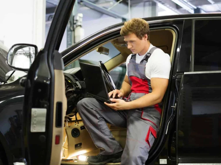 A mechanic in overalls uses a laptop while sitting in the driver's seat of a car with the interior dashboard partially disassembled, conducting a thorough vehicle inspection.