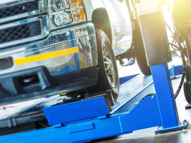A silver pickup truck is positioned on a blue hydraulic lift in an auto repair shop, receiving expert Alignment Services as sunlight shines in from the background.
