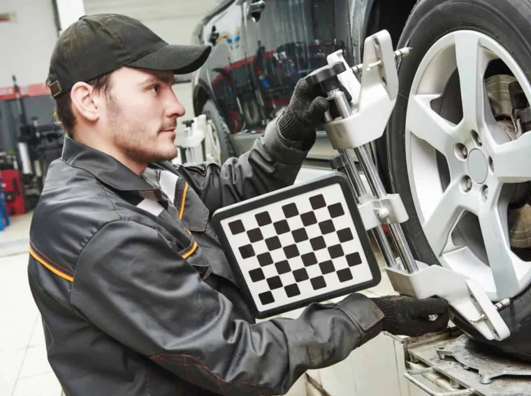 A mechanic uses a wheel alignment tool with a checkered calibration board on a car’s front tire, showcasing the precision of professional Alignment Services in an auto repair shop.