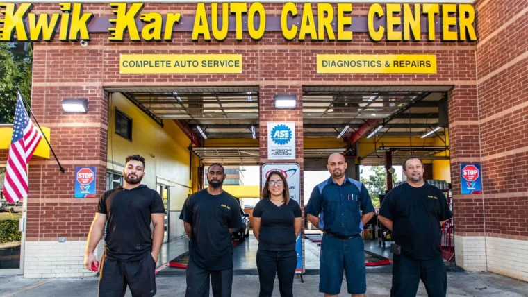Five auto service employees stand in front of the open garage bays at Kwik Kar Auto Care Center, below the shop’s yellow sign.
