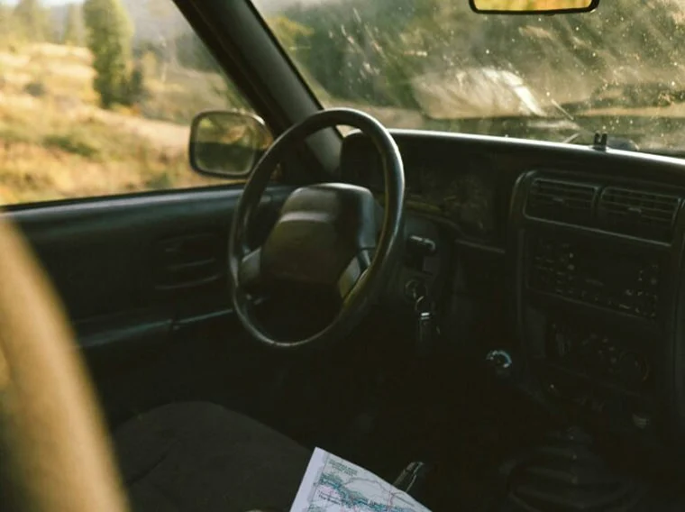 A car interior showing a steering wheel, dashboard, and a map resting on the passenger seat, the scene capturing the character of road trips, with a blurred outdoor landscape visible through the window.