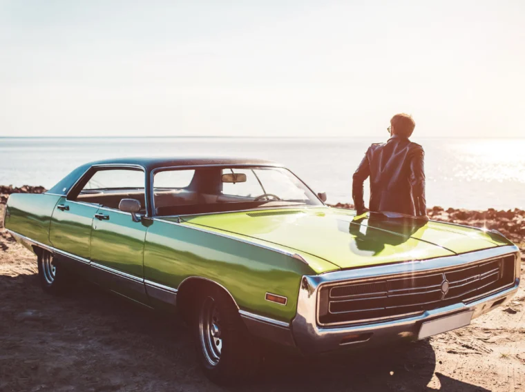 A person in a leather jacket stands by a green vintage car from Cars of Character, parked near a rocky shoreline with sunlight reflecting off the water.