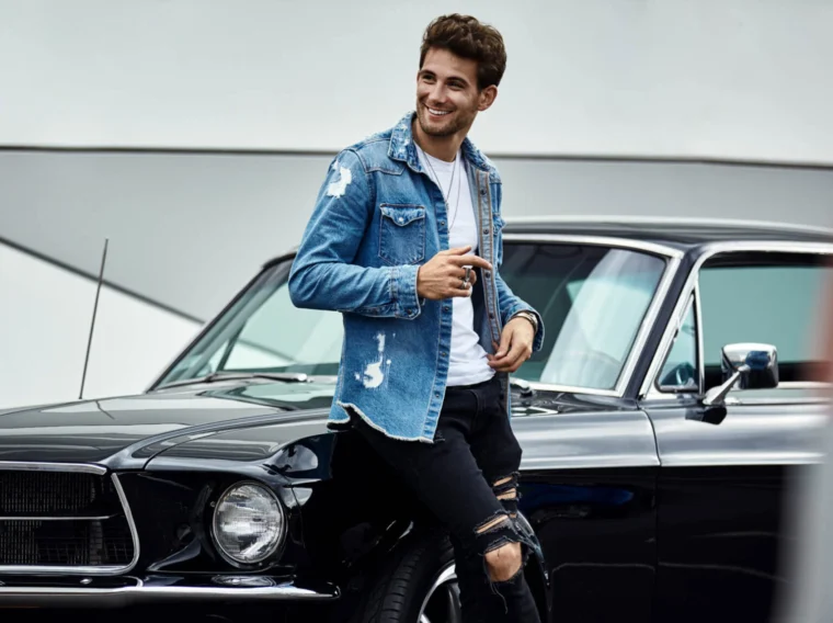 Man wearing a denim jacket and ripped black jeans leans against the front of a black vintage car, smiling and holding sunglasses.