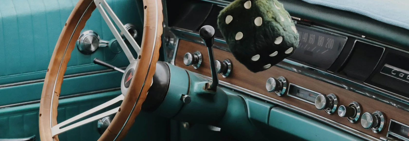 Interior of a vintage car from the Cars of Character collection, featuring a wooden steering wheel, manual gear shift, teal upholstery, and fuzzy dice hanging from the rearview mirror.