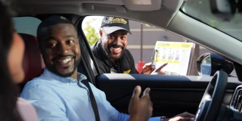 A drive-thru worker smiles and holds a menu beside a car while a man inside the vehicle smiles and gestures toward the passenger.
