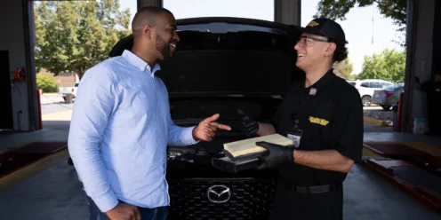 A customer and an employee stand in front of an open car hood at an auto service center, smiling and talking while holding paperwork.