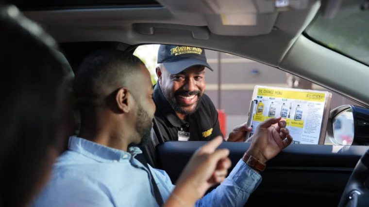 A drive-thru worker hands a menu to two people sitting in a car, as they look at the menu and prepare to order.
