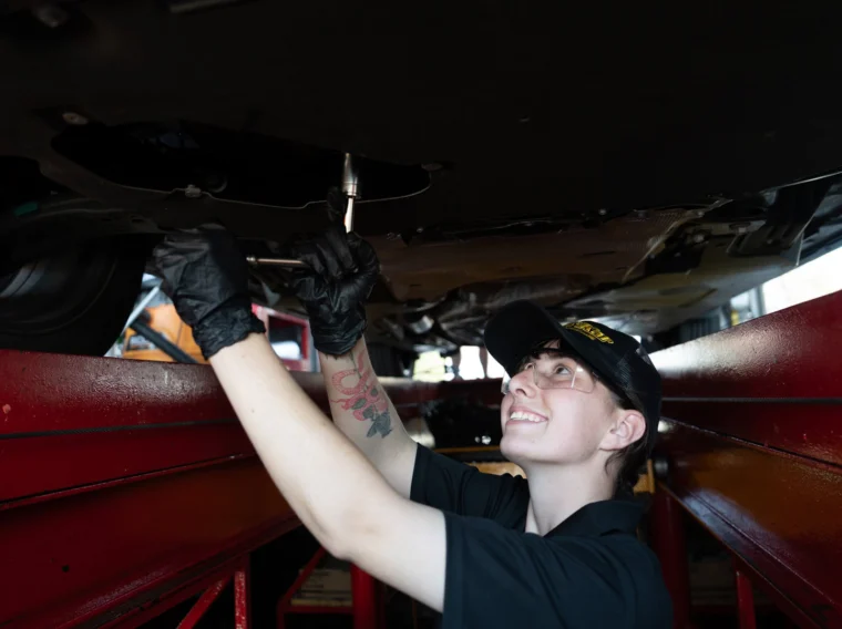 A mechanic wearing gloves and a hat works under a raised vehicle, using a tool to loosen or tighten a part, while smiling.