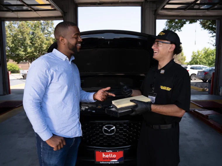 A customer and an auto technician stand in front of a Mazda car with the hood open; the technician holds an air filter and both are smiling.
