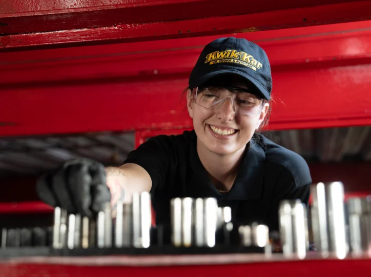 A person wearing safety glasses and a black cap with "AAMCO" logo selects a metal tool from a set inside a red toolbox.